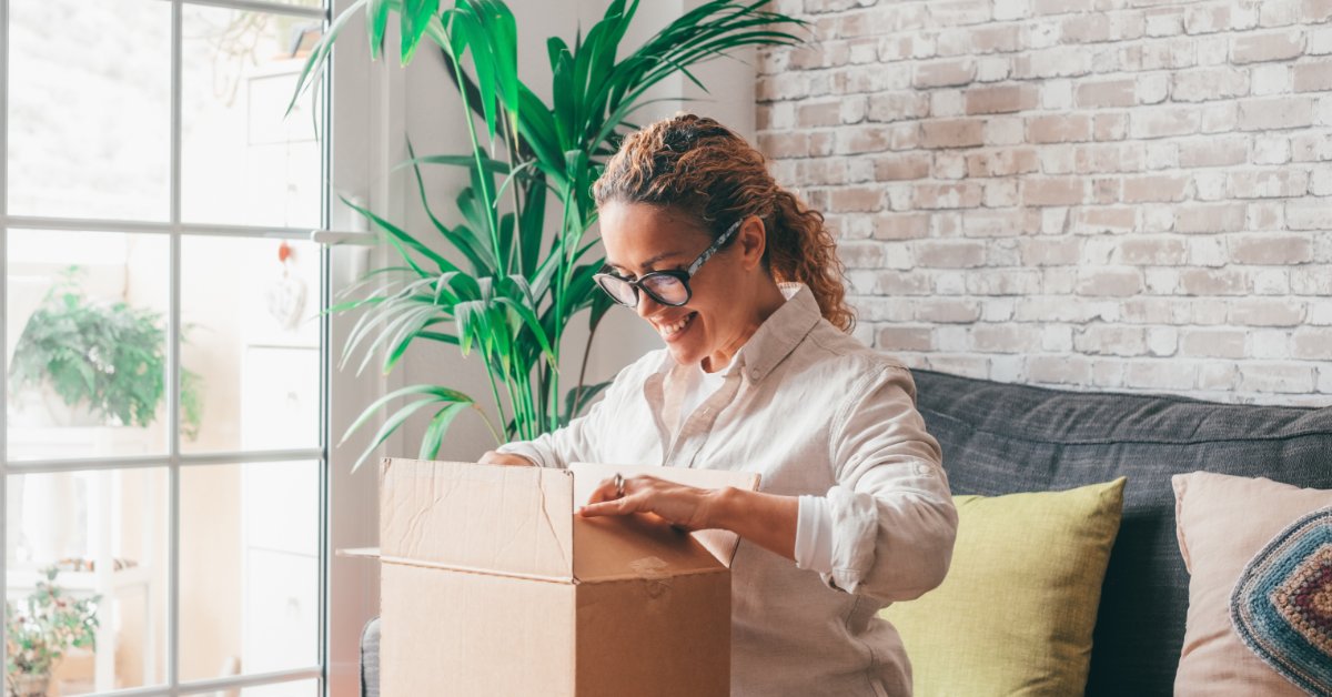 9 Micro Luxury Trends in the Novelty Gift Market A woman wearing glasses opening a cardboard box on a sofa beside pillows, a window, plants, and brick wall.