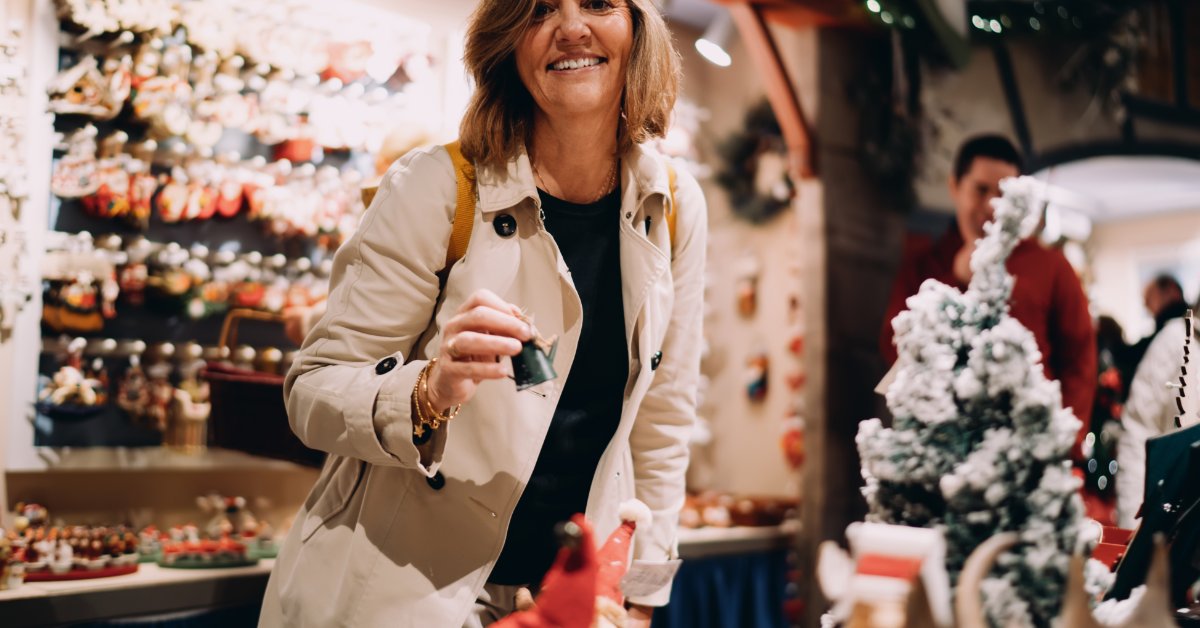 9 Micro Luxury Trends in the Novelty Gift Market A smiling woman in a cream jacket holding a small ornament in a shop filled with holiday decorations.