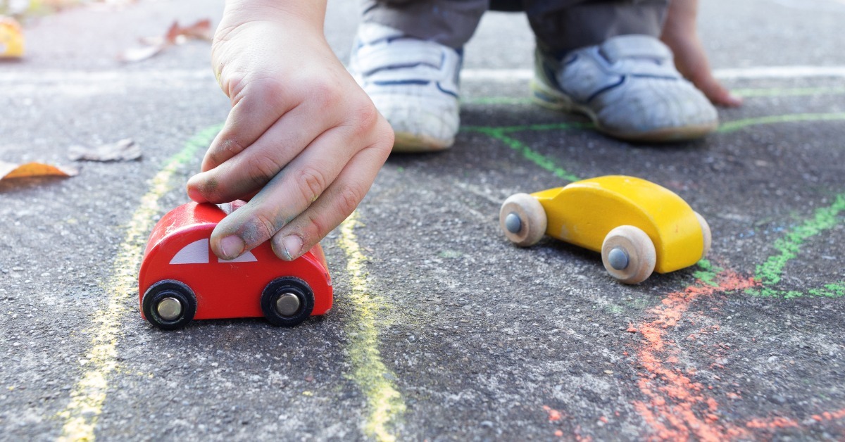 A child pushing a small red wooden toy car across pavement marked with colorful chalk lines beside a yellow toy car.