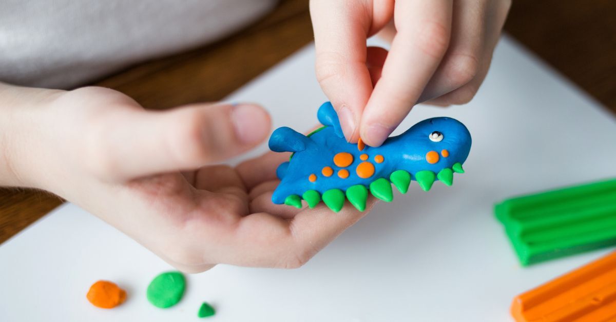 A close-up of a small child creating a blue dinosaur with orange spots and green spikes, all out of clay.