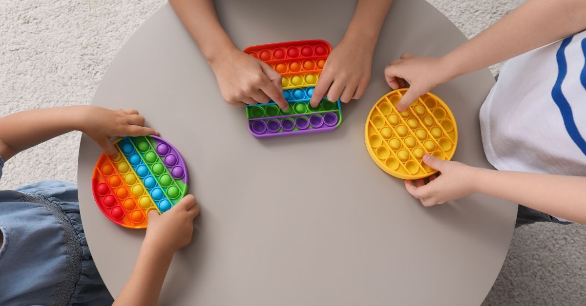 Three young kids sitting around a beige circular table while playing with pop-it fidget toys in various colors.