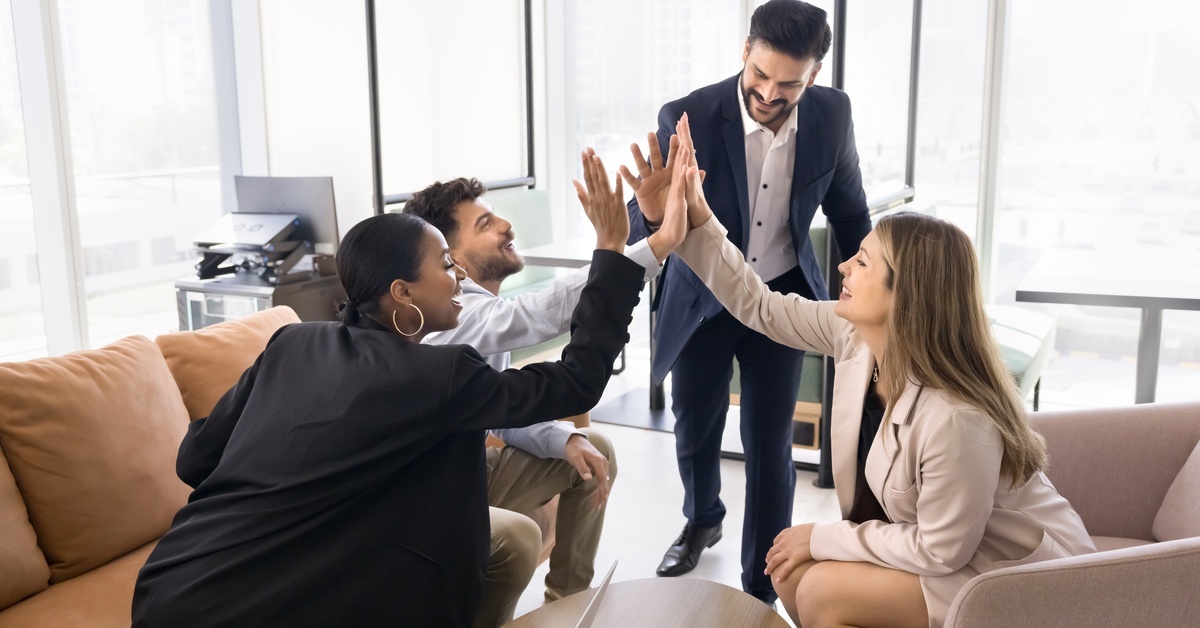Four people in business attire sit on and stand near couches and a chair, high-fiving each other and smiling.