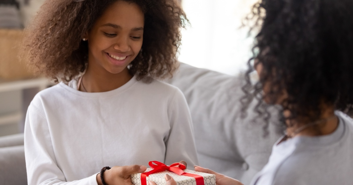Two women in gray shirts sitting on a gray couch exchange a small gift wrapped in giftwrap and tied with a red ribbon. Two women in gray shirts sitting on a gray couch exchange a small gift wrapped in giftwrap and tied with a red ribbon.