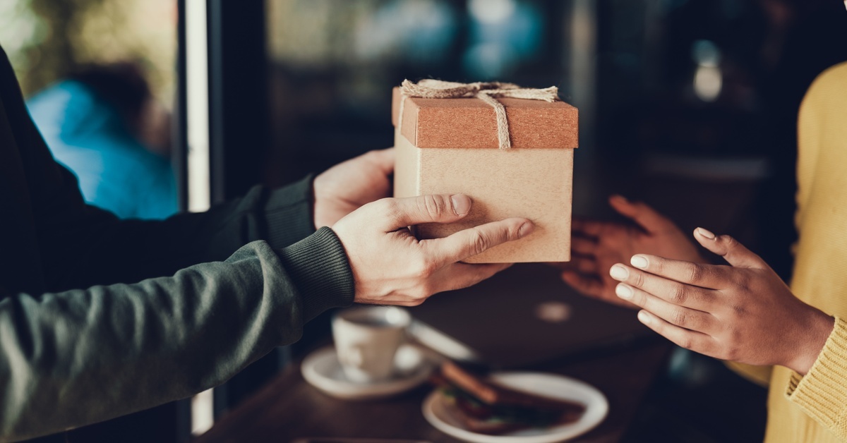 A person hands another person a cardboard giftbox with a tan bow to another person outside a café. A person hands another person a cardboard giftbox with a tan bow to another person outside a café.