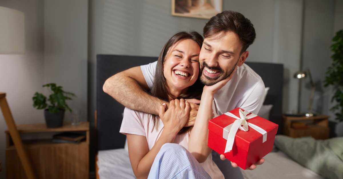 A couple smiles and embraces each other while sitting on the edge of a bed. One holds a gift box.