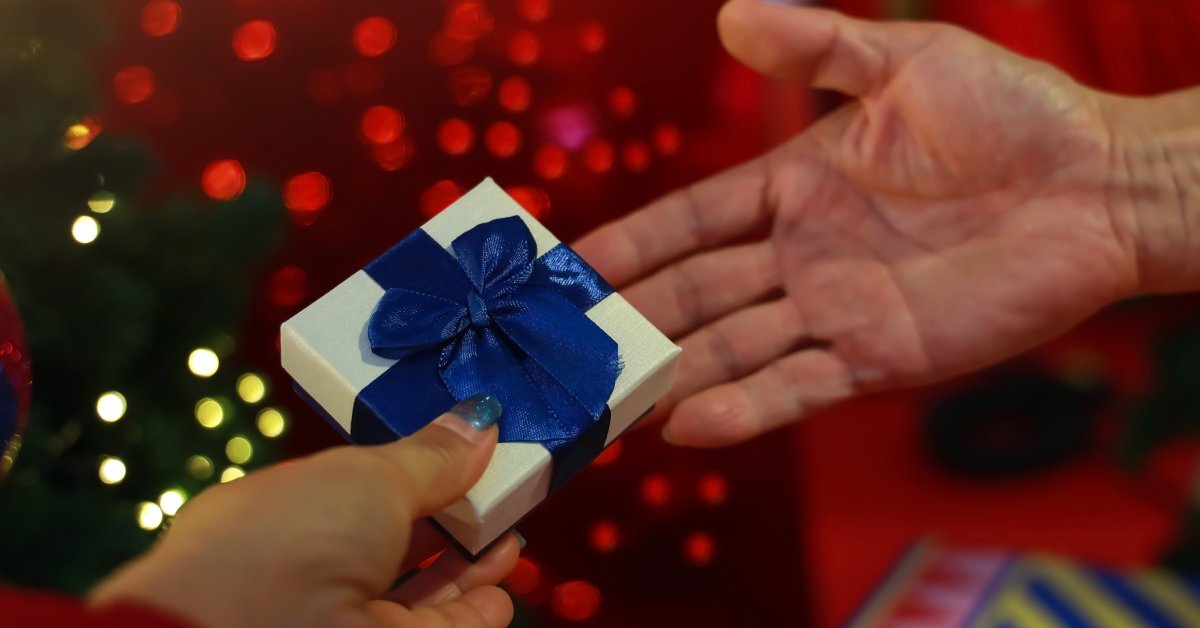 A close up of a hand passing a small, white gift box with a blue bow to another hand in front of shining lights.