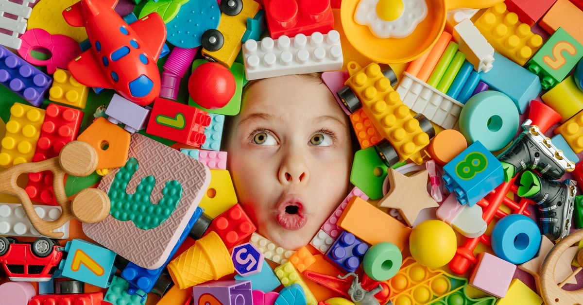 A young child's surprised face emerges from a pile of toys with building blocks, miniature cars, animals, and other objects.
