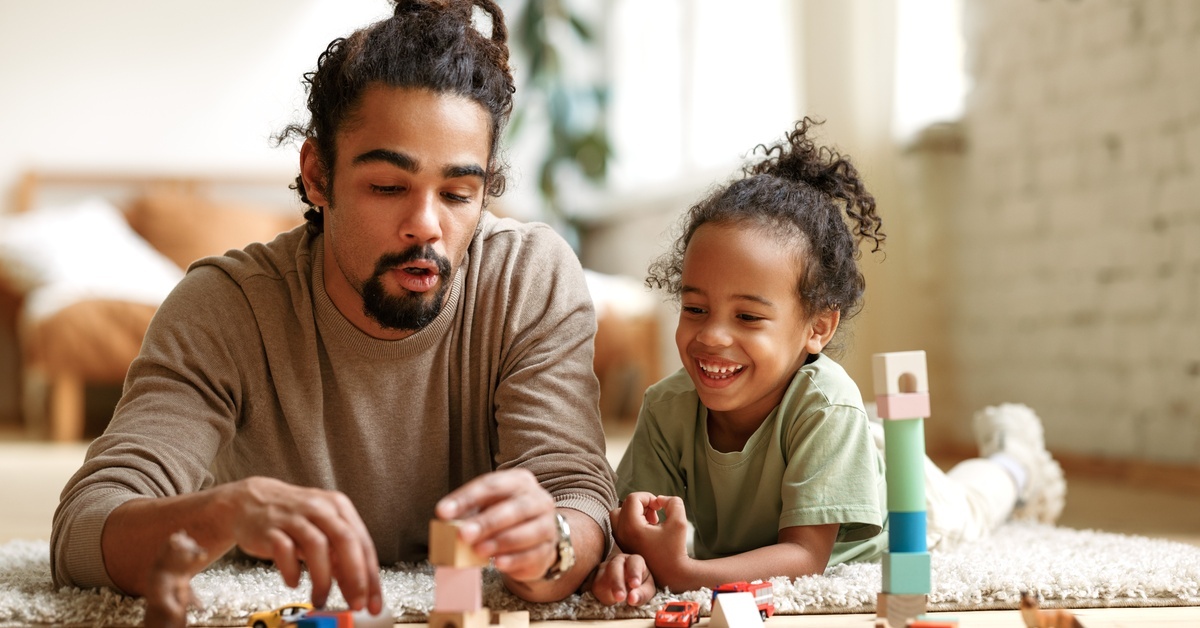 A dad and his child lay on the floor on a carpet, playing with blocks, miniature cars, and animals.