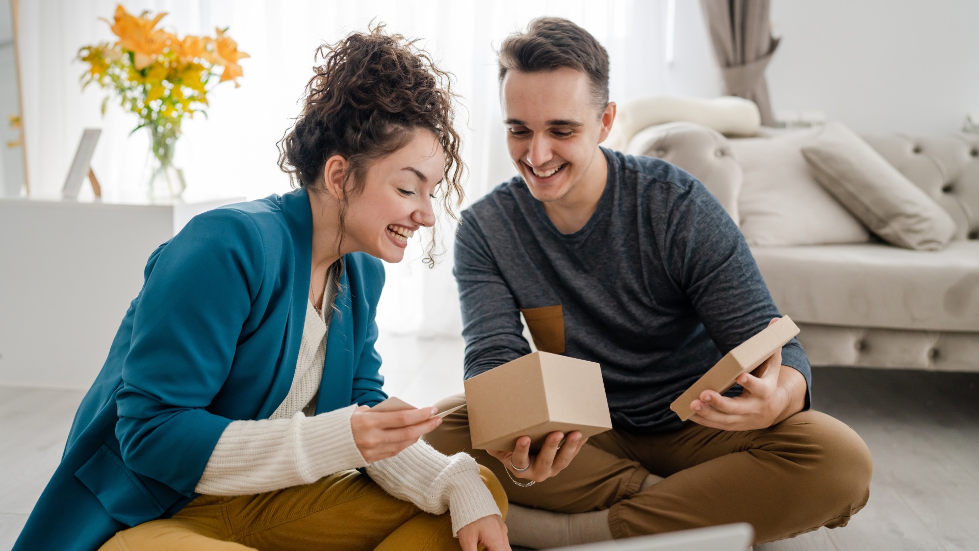 Tiny Gift Ideas for Men That Make a Big Impact A man and a woman sitting on the floor, opening a gift box and smiling wide while looking inside.