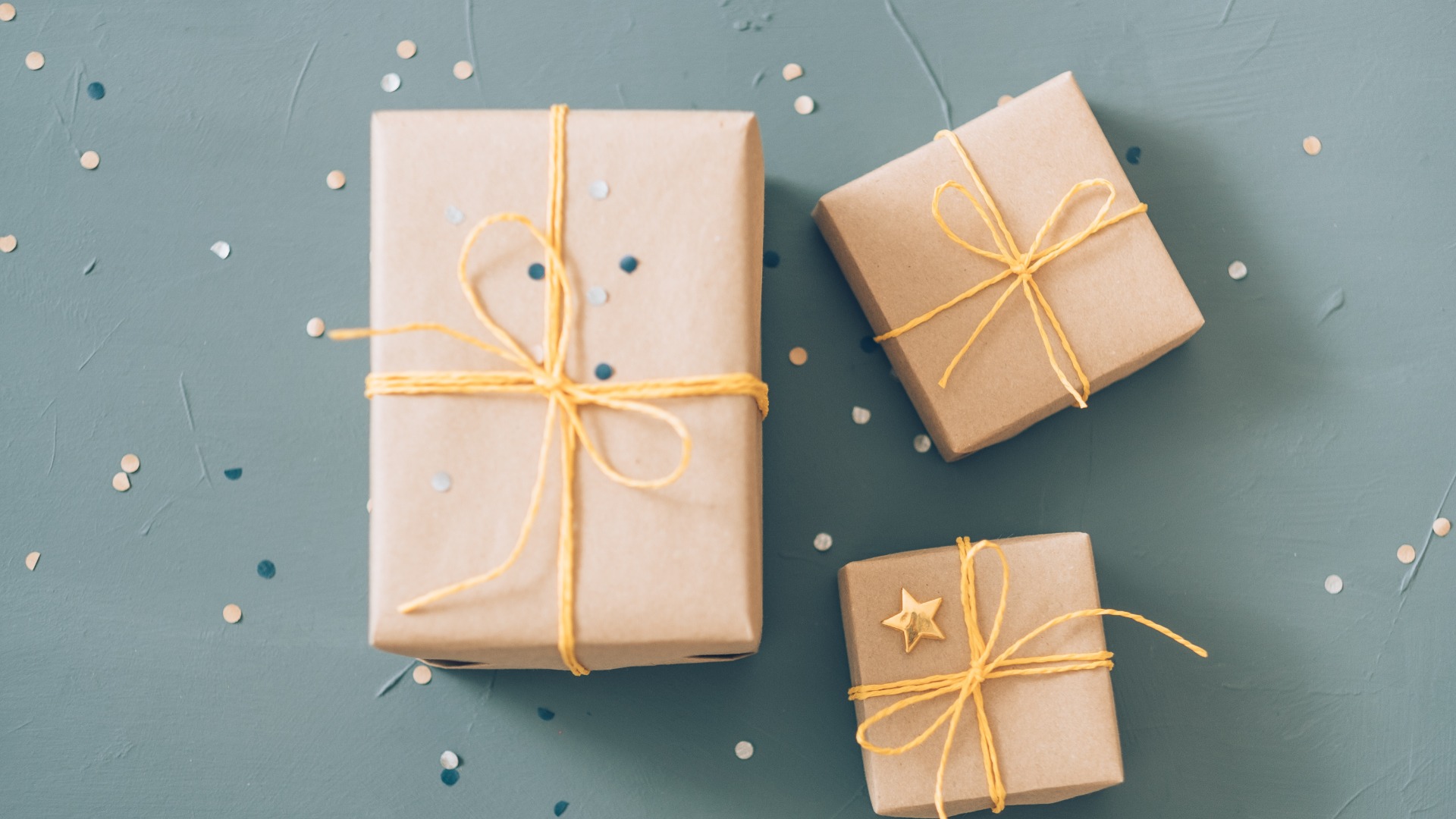 Tiny Gift Ideas for Men That Make a Big Impact A top-down view of a pile of gifts wrapped in brown paper amongst small, glitter pieces on a surface.