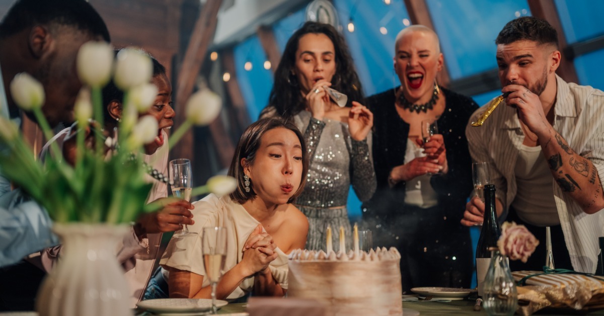 A group of young adults standing around a table with party horns in their hands, watching a girl blow out candles on a cake.