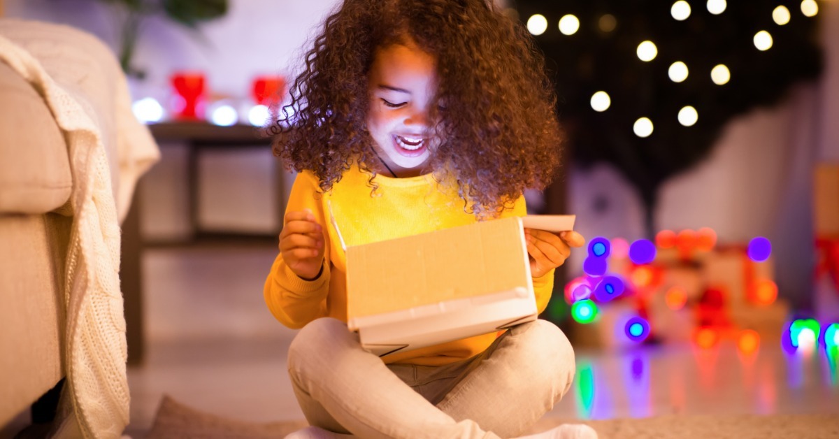 a child sitting on a rug and opening a glowing box near colorful lights and a dark holiday tree indoors.