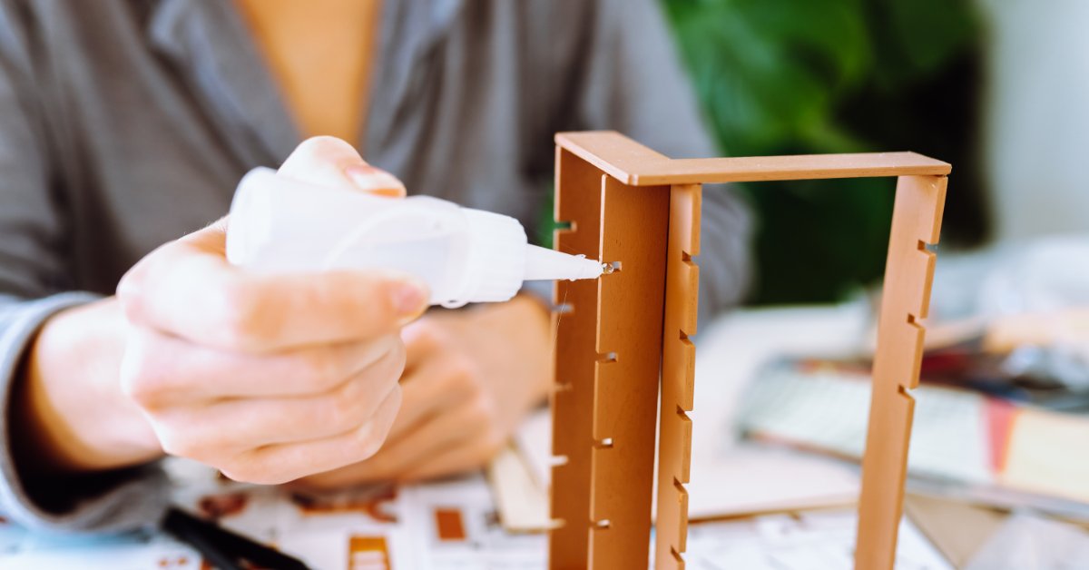 Tiny Architecture Models for Creative Adults Hands applying glue while assembling a wooden house model on a table with small craft pieces and plants nearby.